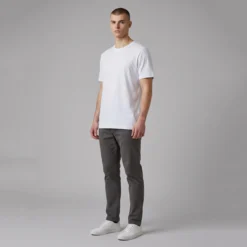 Man standing wearing a plain white t-shirt, dark gray trousers, and white sneakers against a gray background