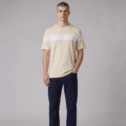 Man wearing a beige crewneck t-shirt with a white horizontal chest stripe and dark blue jeans